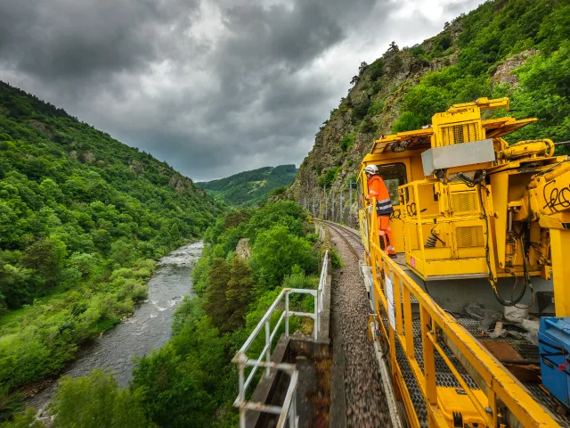 Engin de maintenance ferroviaire circulant dans une vallée encaissée, avec un opérateur en intervention.