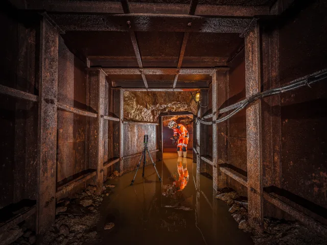 Un homme en combinaison orange dans la mine de la Rodde avec de l'eau au sol et un trépied.