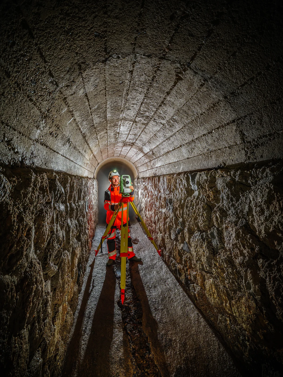 Ouvrier en tenue de sécurité utilisant une station totale dans un tunnel en pierre.
