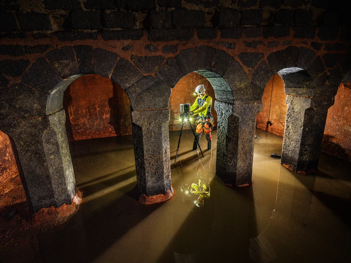 Technicien dans un environnement souterrain utilisant un appareil de mesure au-dessus de l’eau stagnante dans un espace avec des arches en pierre.