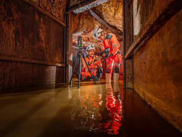 Deux personnes en tenue de sécurité orange analysent des données dans un tunnel inondé, entourées de murs en métal.