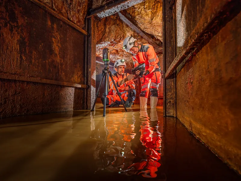Deux personnes en tenue de sécurité orange analysent des données dans un tunnel inondé, entourées de murs en métal.