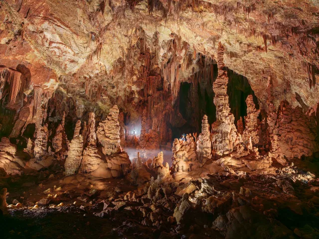 Intérieur d'une grotte avec des stalagmites, éclairage mettant en valeur les formations rocheuses et une silhouette humaine au fond.