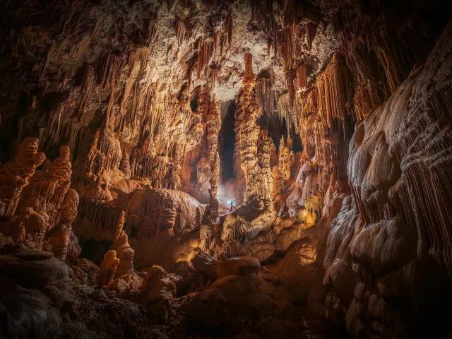 Caverne souterraine avec formations de stalagmites et stalactites, explorateur en bleu éclairé par une lumière.