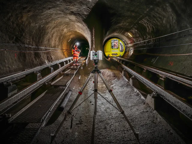 Une personne en tenue de sécurité dans un tunnel avec des rails et un train visible à droite.