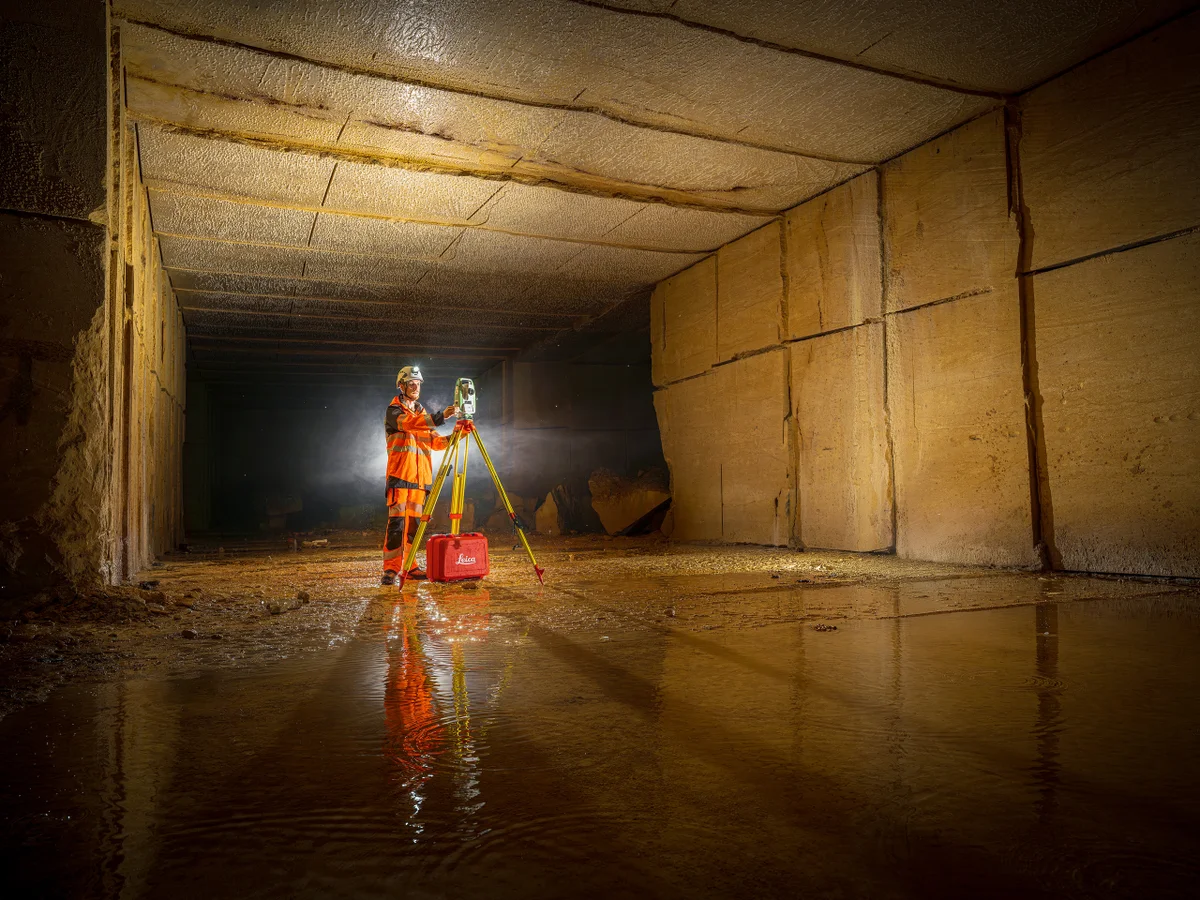 Un homme en tenue orange utilisant un appareil de mesure dans un environnement sombre et humide, reflet sur le sol.