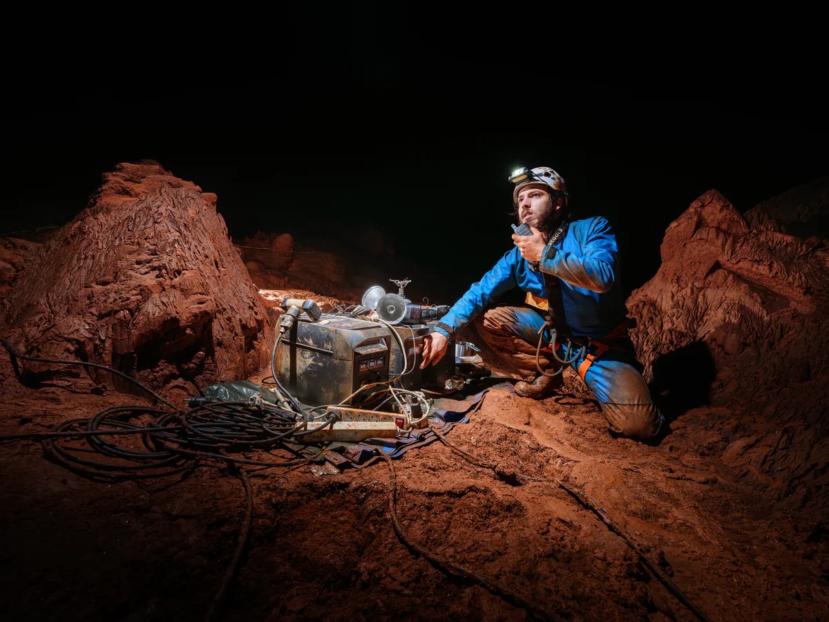 Spéléologue utilisant un équipement dans une grotte sombre avec un sol en terre rouge.