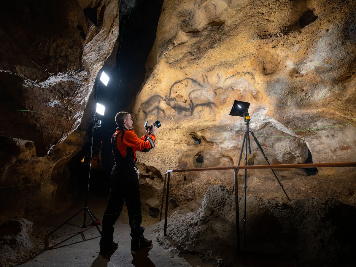 Homme photographiant des peintures rupestres dans une caverne éclairée par des lampes.