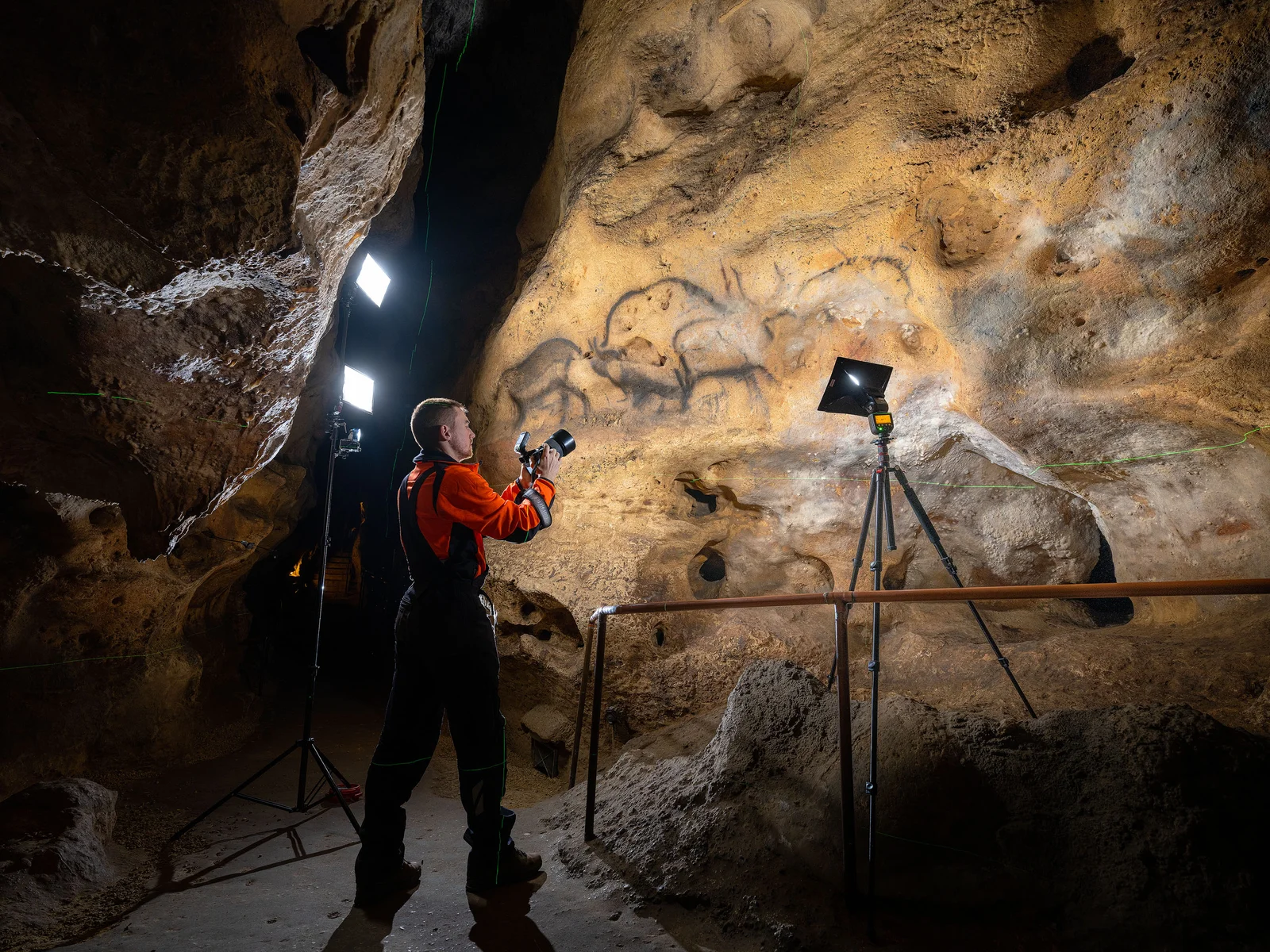 Homme photographiant des peintures rupestres dans une caverne éclairée par des lampes.