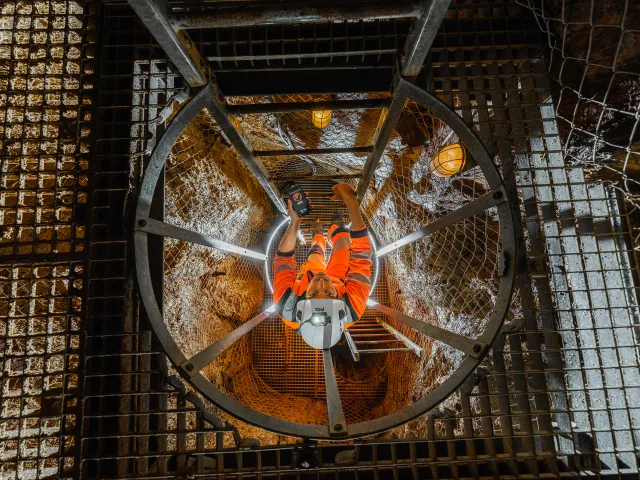 Une personne en tenue orange descend une échelle dans un puits métallique, tenant un appareil de mesure.