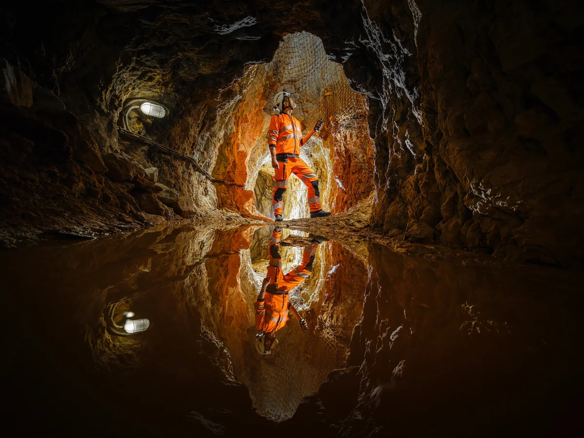 Mineur dans un tunnel souterrain avec des murs de roche et de l’eau stagnante.
